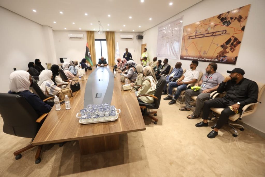 Group of men and women seated around a long conference table in a meeting room, with a Libyan flag and a banner on the wall behind them.