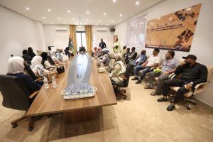 Group of men and women seated around a long conference table in a meeting room, with a Libyan flag and a banner on the wall behind them.