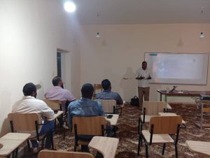 28B95899-8E56-47C2-98F5-86390EEFAF0E - جامعة سبها | Sebha University Teacher giving a presentation at the front of a classroom with students seated at desks facing him.