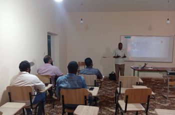 Teacher giving a presentation at the front of a classroom with students seated at desks facing him.