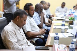 294F58B1-5A12-47ED-B12B-59A3ADBFF9EB - جامعة سبها | Sebha University Group of men seated around a conference table, the foreground man in a striped shirt looking down, with papers and water bottles on the table.