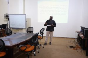 2A500850-995F-485F-A226-D09EA5E8BF49 - جامعة سبها | Sebha University Man giving a presentation in a conference room, projected Arabic text on the wall behind him, holding papers and gesturing.