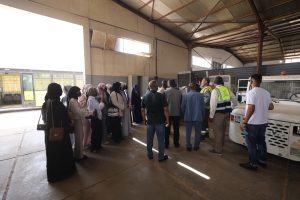 Group of people in a large indoor facility listening to staff in safety vests beside a parked bus, likely a briefing or tour.
