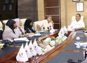 People seated around a conference table in a meeting room, several women wearing hijabs and men talking, with drinks and papers on the table and small flag banners in front of the participants.