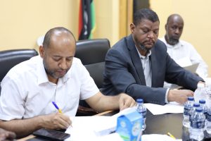 2D88DC4F-FA48-4F2A-A61A-D02E9BF47DB0 - جامعة سبها | Sebha University Three men sit at a conference table signing or reviewing papers during a meeting, with water bottles and documents in view.