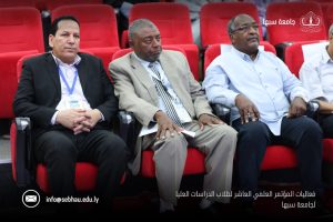 2DF68F02-1E65-4FBE-8B90-A06FBDE85DC5 - جامعة سبها | Sebha University Three men in suits sit in red upholstered conference seating, wearing badges at a formal event.",
