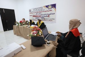 Panel of graduates in robes seated at a long table with a floral centerpiece and banner behind, in a bright room