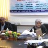 Two adults seated at a conference table with papers, a laptop, and a floral centerpiece, a banner in Arabic on the wall behind them.