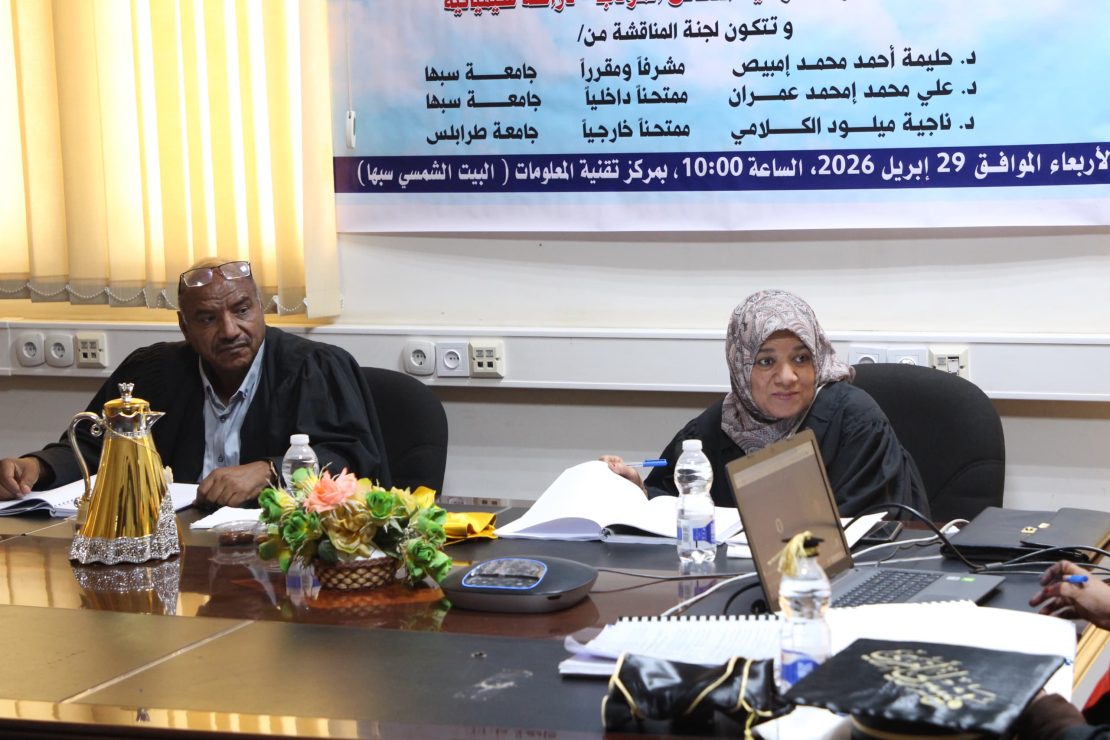 Two adults seated at a conference table with papers, a laptop, and a floral centerpiece, a banner in Arabic on the wall behind them.