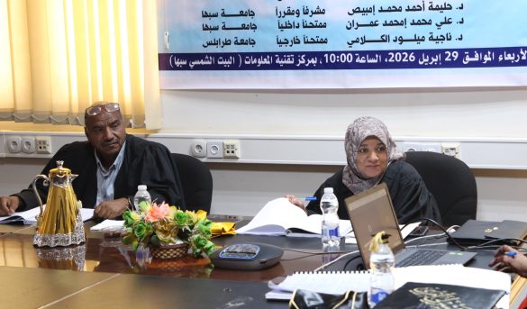 Two adults seated at a conference table with papers, a laptop, and a floral centerpiece, a banner in Arabic on the wall behind them.