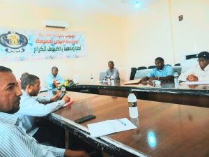 Group of men seated around a long conference table in a meeting room, banner with Arabic text on the wall behind them.