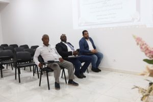 3AA410B1-229F-47C9-920E-8A153AADCF79 - جامعة سبها | Sebha University Three men sit in the front row of a conference room, paying attention to a projected presentation on the wall behind them.