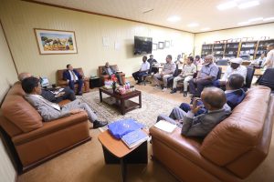 3B327264-E197-465A-9846-B41328DFF0E4 - جامعة سبها | Sebha University Group of people seated in a U-shaped arrangement in a beige-office meeting room with a coffee table and snacks.