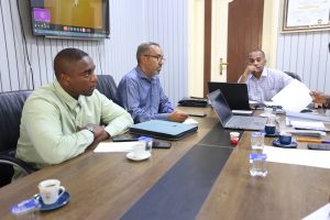 Three men sit at a conference table with laptops, papers, and coffee cups, engaged in a meeting.