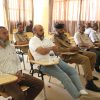 Group of men seated in a classroom, several in tan uniforms, listening at small desks with water bottles nearby.