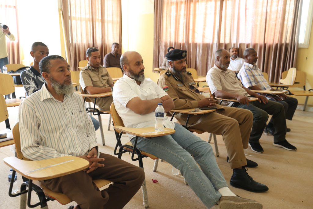 3FC844AF-494E-4D7B-ADFE-AACD768C1192 - جامعة سبها | Sebha University Group of men seated in a classroom, several in tan uniforms, listening at small desks with water bottles nearby.