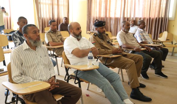3FC844AF-494E-4D7B-ADFE-AACD768C1192 - جامعة سبها | Sebha University Group of men seated in a classroom, several in tan uniforms, listening at small desks with water bottles nearby.