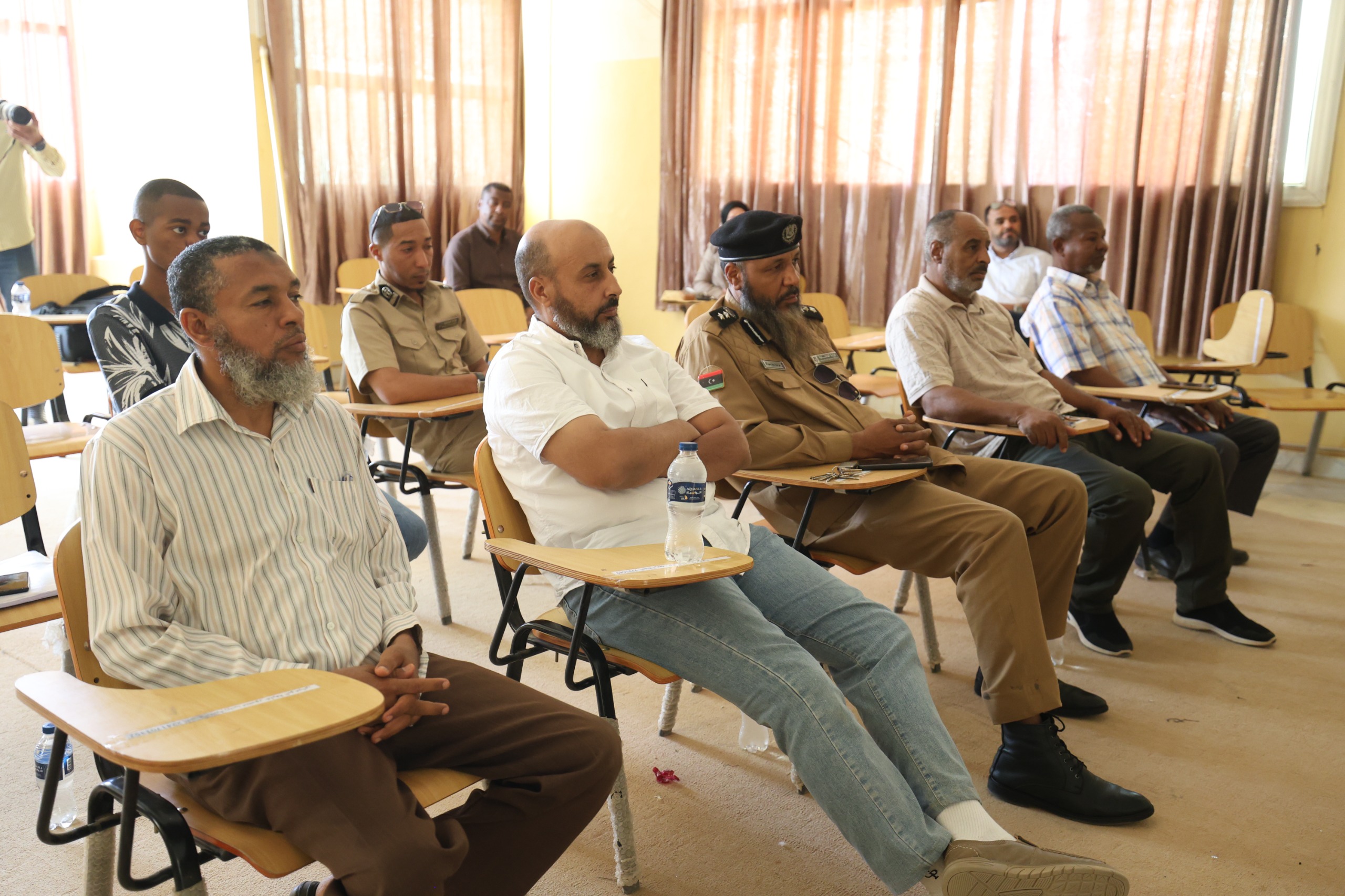Group of men seated in a classroom, several in tan uniforms, listening at small desks with water bottles nearby.