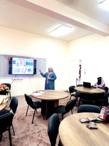 Woman in a headscarf points at a slide on a wall-mounted screen during a presentation in a classroom; another person sits at a laptop nearby.