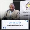 Man in a beige suit delivering a speech at a podium with a microphone, a banner with Arabic text in the background behind him.