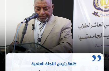 Man in a beige suit delivering a speech at a podium with a microphone, a banner with Arabic text in the background behind him.