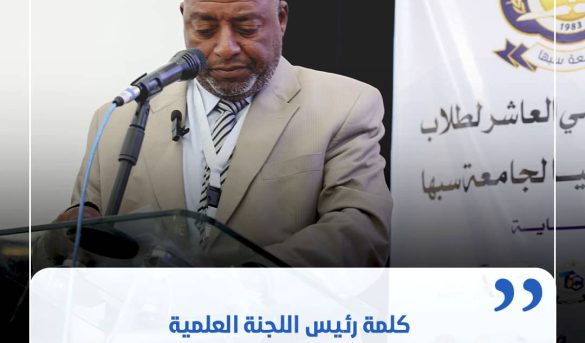 48C00A05-D559-4110-B1D6-77842360E751 - جامعة سبها | Sebha University Man in a beige suit delivering a speech at a podium with a microphone, a banner with Arabic text in the background behind him.