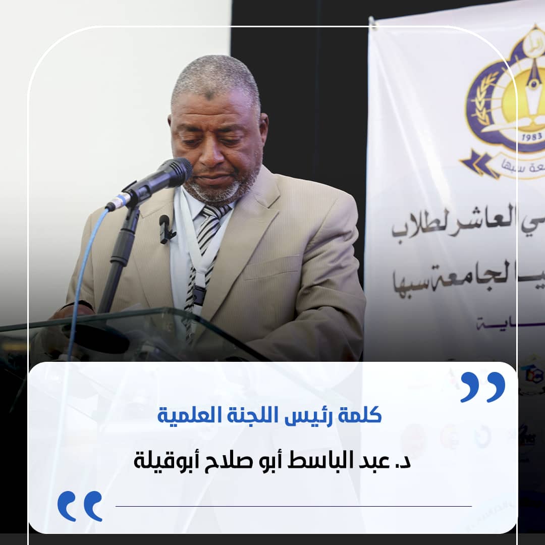 Man in a beige suit delivering a speech at a podium with a microphone, a banner with Arabic text in the background behind him.