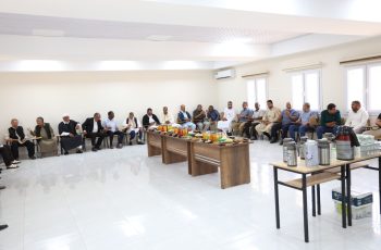 Group of men seated along walls in a bright conference room, with snacks and drinks on a central table in front of them.