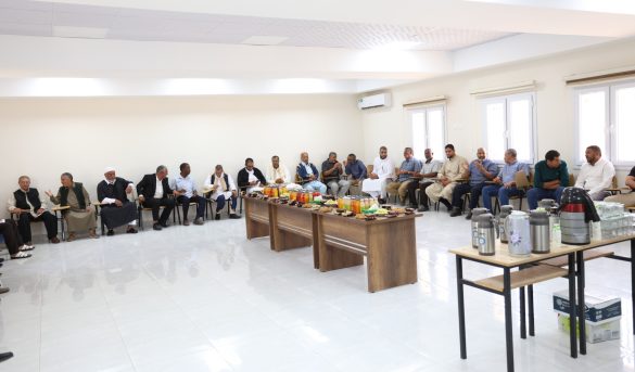 494ED8E0-F76E-408E-AD22-57C3729A502B - جامعة سبها | Sebha University Group of men seated along walls in a bright conference room, with snacks and drinks on a central table in front of them.