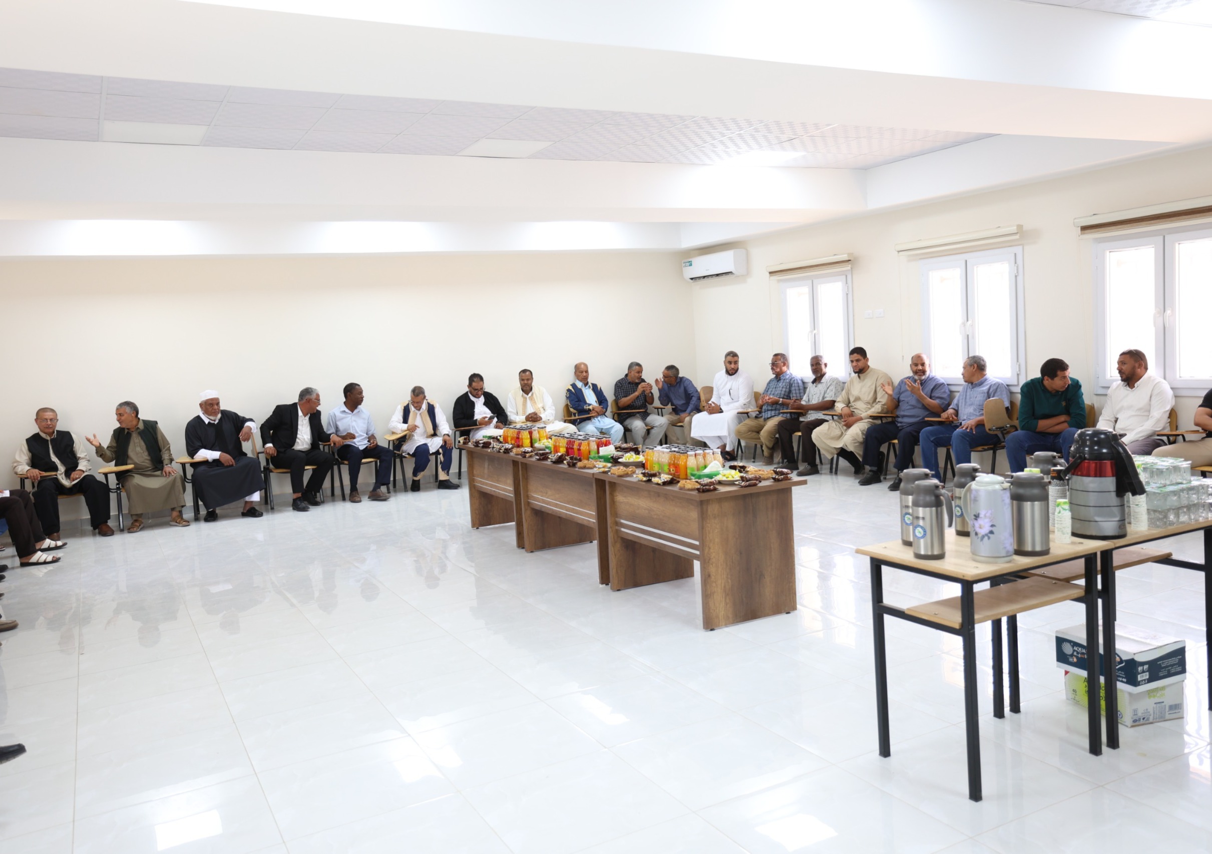 Group of men seated along walls in a bright conference room, with snacks and drinks on a central table in front of them.