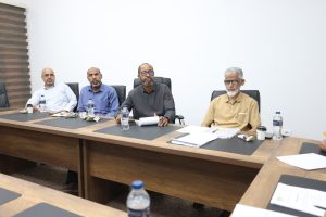 4B6AE31C-6BA4-454A-8BC0-FDE48CCA2933 - جامعة سبها | Sebha University Four men sit at a conference table during a meeting, with papers, notebooks and water bottles in front of them.