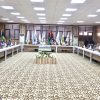 People sit around a U-shaped conference table in a formal meeting room with flags lined up along the back wall and a central flower arrangement on a low podium.