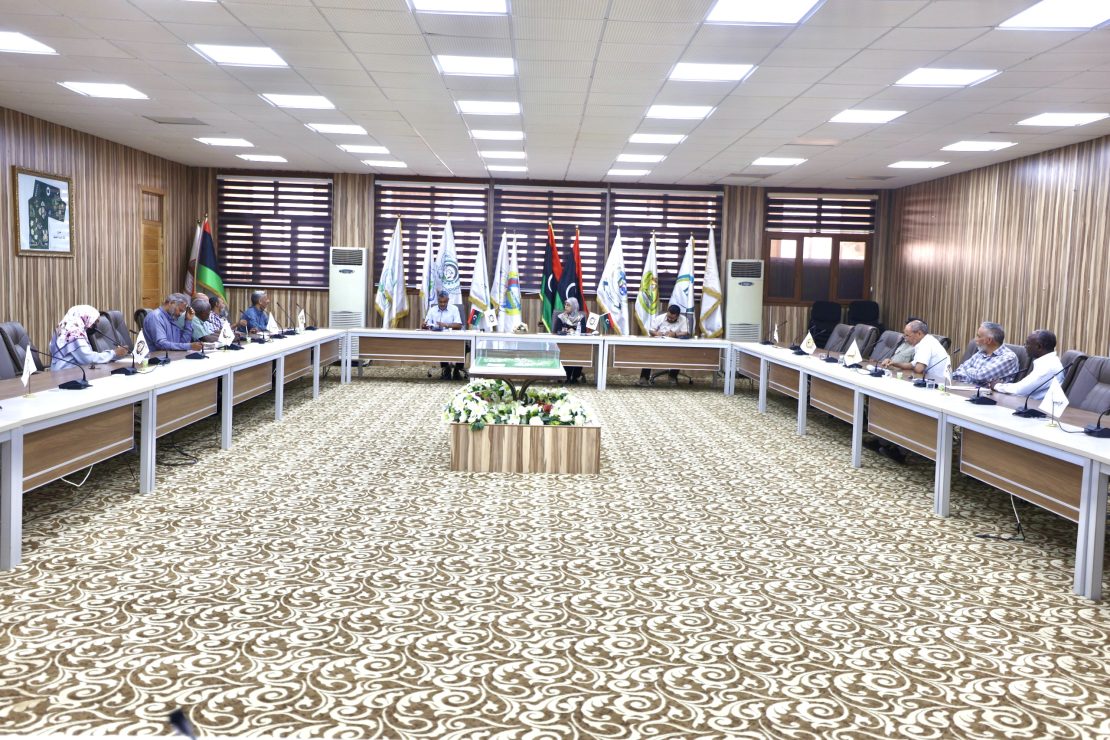 People sit around a U-shaped conference table in a formal meeting room with flags lined up along the back wall and a central flower arrangement on a low podium.