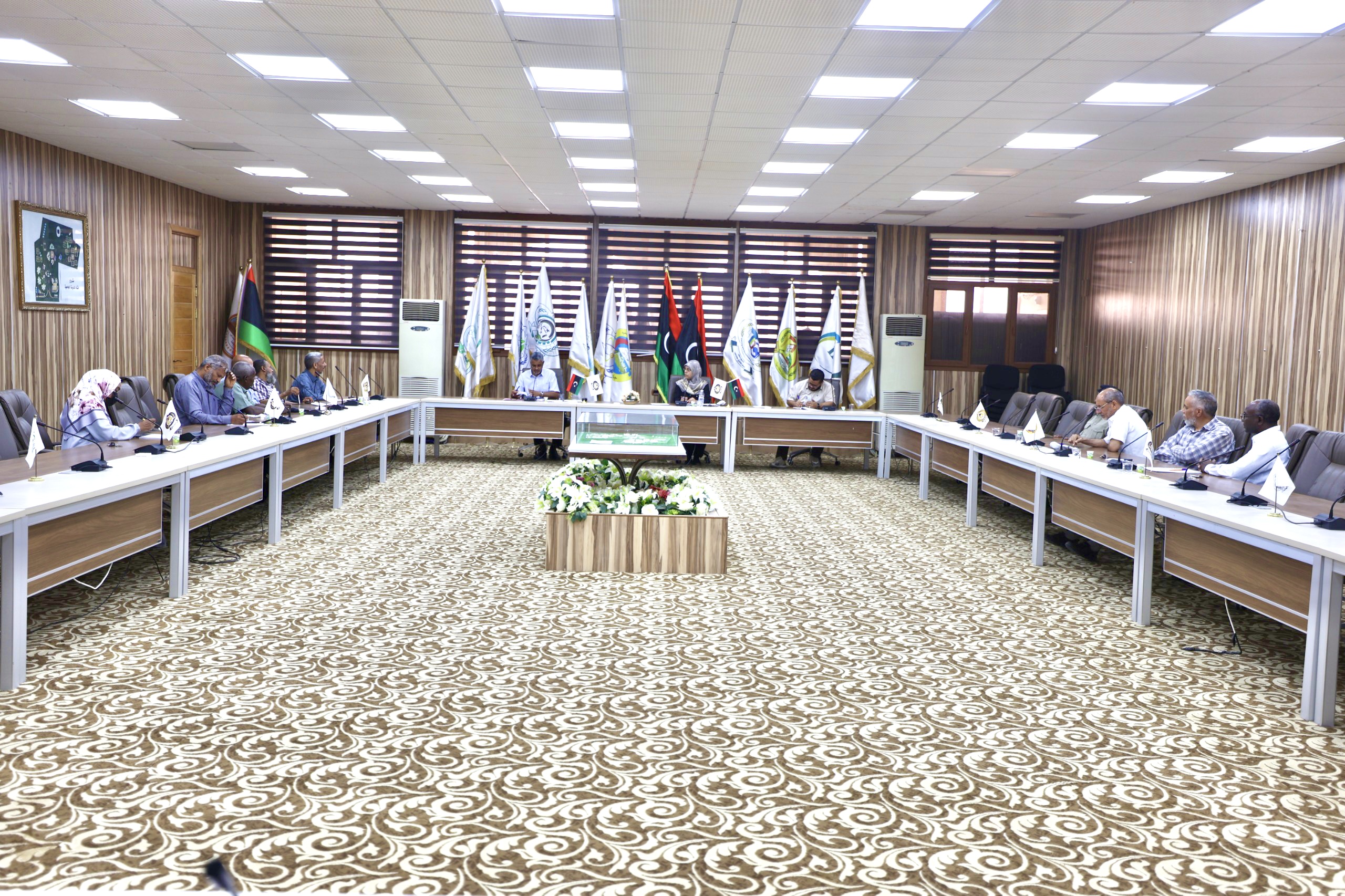 People sit around a U-shaped conference table in a formal meeting room with flags lined up along the back wall and a central flower arrangement on a low podium.