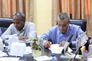 4D2511E1-7FDA-40EE-B9F7-E8C4C784C24D - جامعة سبها | Sebha University Two men sit at a conference table, writing and reviewing documents in a busy office setting with bottles and plants on the table