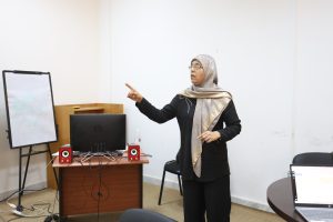 4DE0F812-3853-4159-B5C8-B48DC27C1754 - جامعة سبها | Sebha University Woman in a patterned beige hijab presents at a desk with a computer and red speakers in a classroom.2) Presenter wearing glasses and a hijab points while giving a lecture in a small classroom.3) Classroom setup with a standing instructor, desk, computer, cables, and whiteboard on the left.