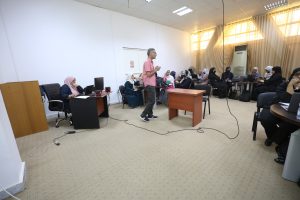 4EF36658-CCA8-4DE5-B319-4925E850ADF5 - جامعة سبها | Sebha University A man in a pink shirt stands at a small wooden podium addressing a classroom full of women wearing hijabs, all with laptops. The room has beige carpet, white walls, and large windows with sheer curtains in the background.