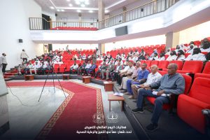 4U1A8827 - جامعة سبها | Sebha University Audience seated in a large auditorium with red seats during a conference or ceremony, some people wearing masks and white coats, a tripod near the stage.