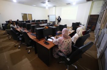 Instructor presenting at the front of a computer lab as students sit around a U-shaped desk setup, viewing a projection on the wall.