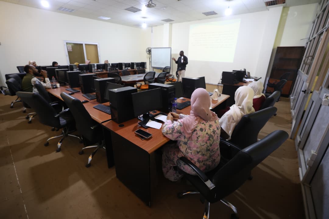 Instructor presenting at the front of a computer lab as students sit around a U-shaped desk setup, viewing a projection on the wall.