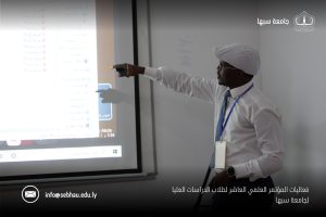 5361DE75-B445-45B2-B0AE-670C408D9F6E - جامعة سبها | Sebha University Man in white shirt and light blue tie presenting with a projected screen to the side; conference branding and contact email appear at the bottom overlay.