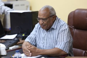 5435F1EC-B76D-4ADF-8C9C-38D0621F716B - جامعة سبها | Sebha University Older man with gray hair and glasses sits at a desk in an office, hands folded over papers, with computer and printer in the background.