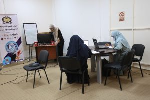 57CEADEA-499E-4C2C-8BBC-5E1F47DF1527 - جامعة سبها | Sebha University Three women in hijabs working in a classroom-like office; one at a computer station near a banner, two at a round table with laptops.