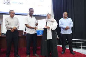 586BF895-FA35-4461-9152-D2EA204CAD19 - جامعة سبها | Sebha University Woman in hijab receives a certificate from a man on stage while two other men stand nearby, a projector screen behind them and a red carpet underfoot.