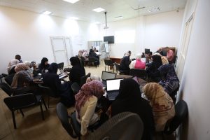 5A02A80E-5559-49E7-8C37-C37FAE48F3F3 - جامعة سبها | Sebha University Group of people, mostly women in hijabs, seated around tables with laptops in a bright classroom or meeting room, projector screen at front.
