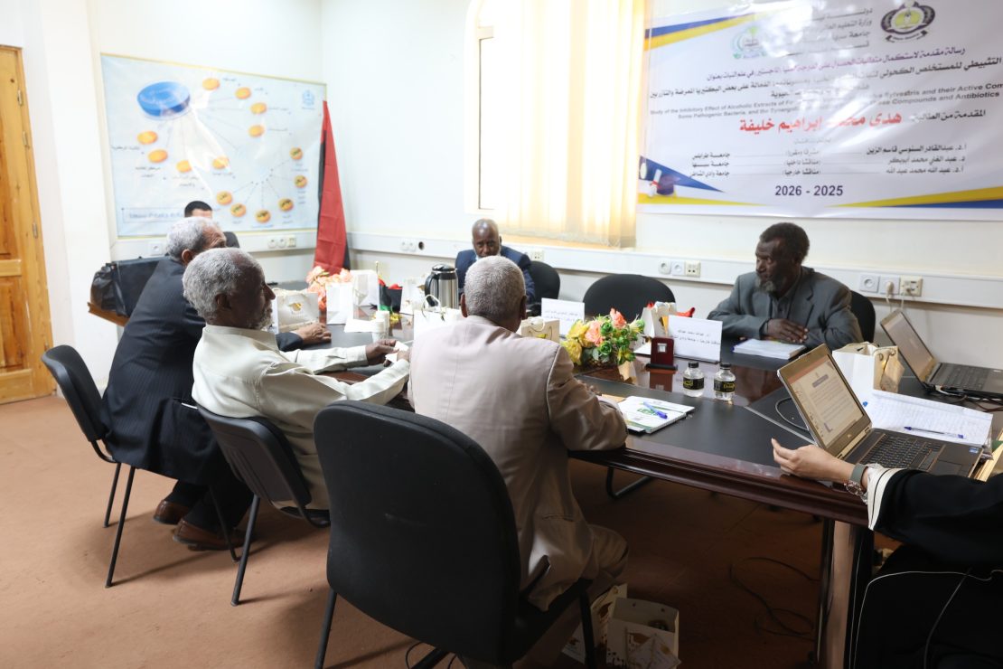 Group of men seated around a conference table in a meeting room, with laptops, paperwork, and gift bags on the table, banner on the wall behind them.