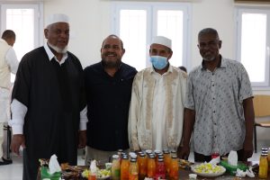 Four men stand together behind a table of drinks and snacks at an indoor gathering; one man wears a face mask.