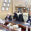 Group of professionals seated around a conference table, discussing in a room with framed certificates and flags on the wall behind them.