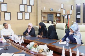 Group of professionals seated around a conference table, discussing in a room with framed certificates and flags on the wall behind them.