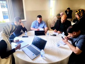 Group of people seated around a round table in a meeting, with laptops, papers, and bottled water visible.